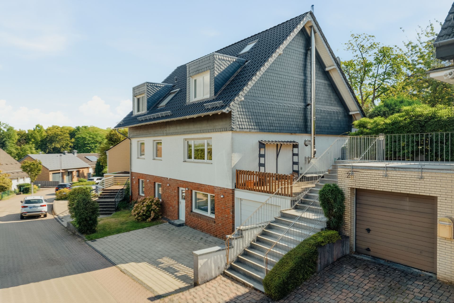 Zweistöckiges Wohnhaus mit Dachgeschoss, Außentreppe und Garage auf gepflasterter Einfahrt.