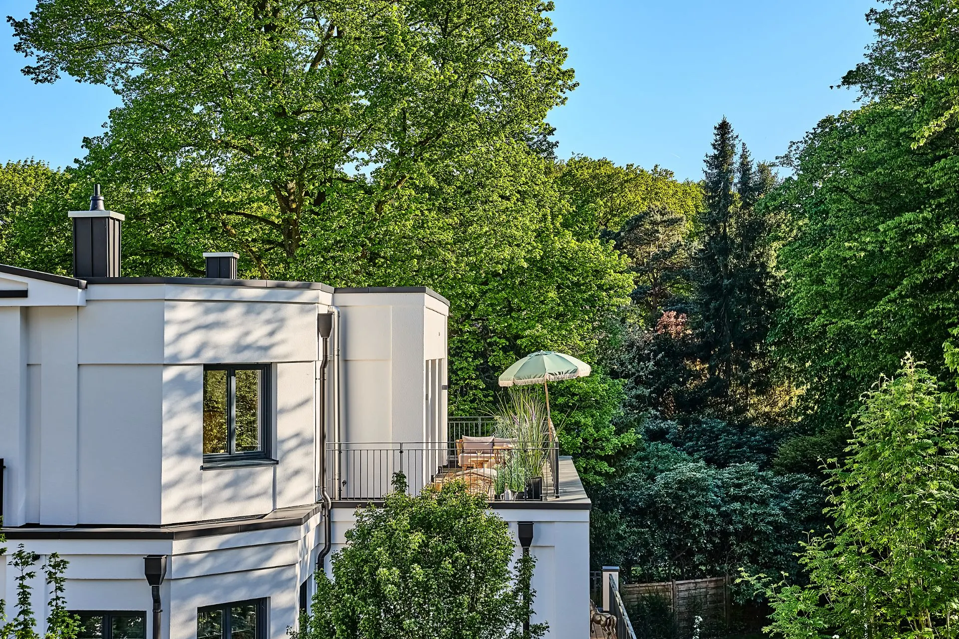 Moderne Hausfassade mit Balkon und Sonnenschirm, umgeben von vielen Bäumen.