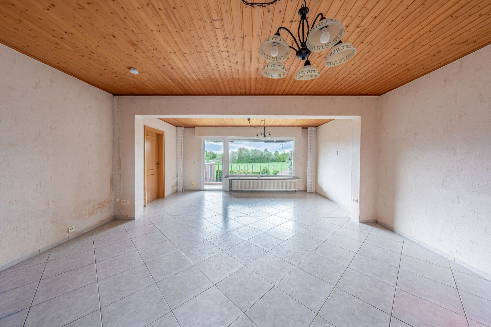 Leeres Wohnzimmer mit Fliesenboden, Holzdecke und großen Fenstern mit Blick ins Grüne.