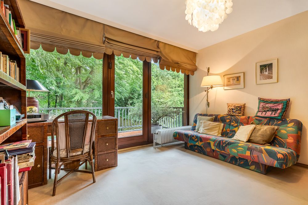 Helles Wohnzimmer mit bunter Couch, Schreibtisch, Regal und großer Fensterfront zum Garten.