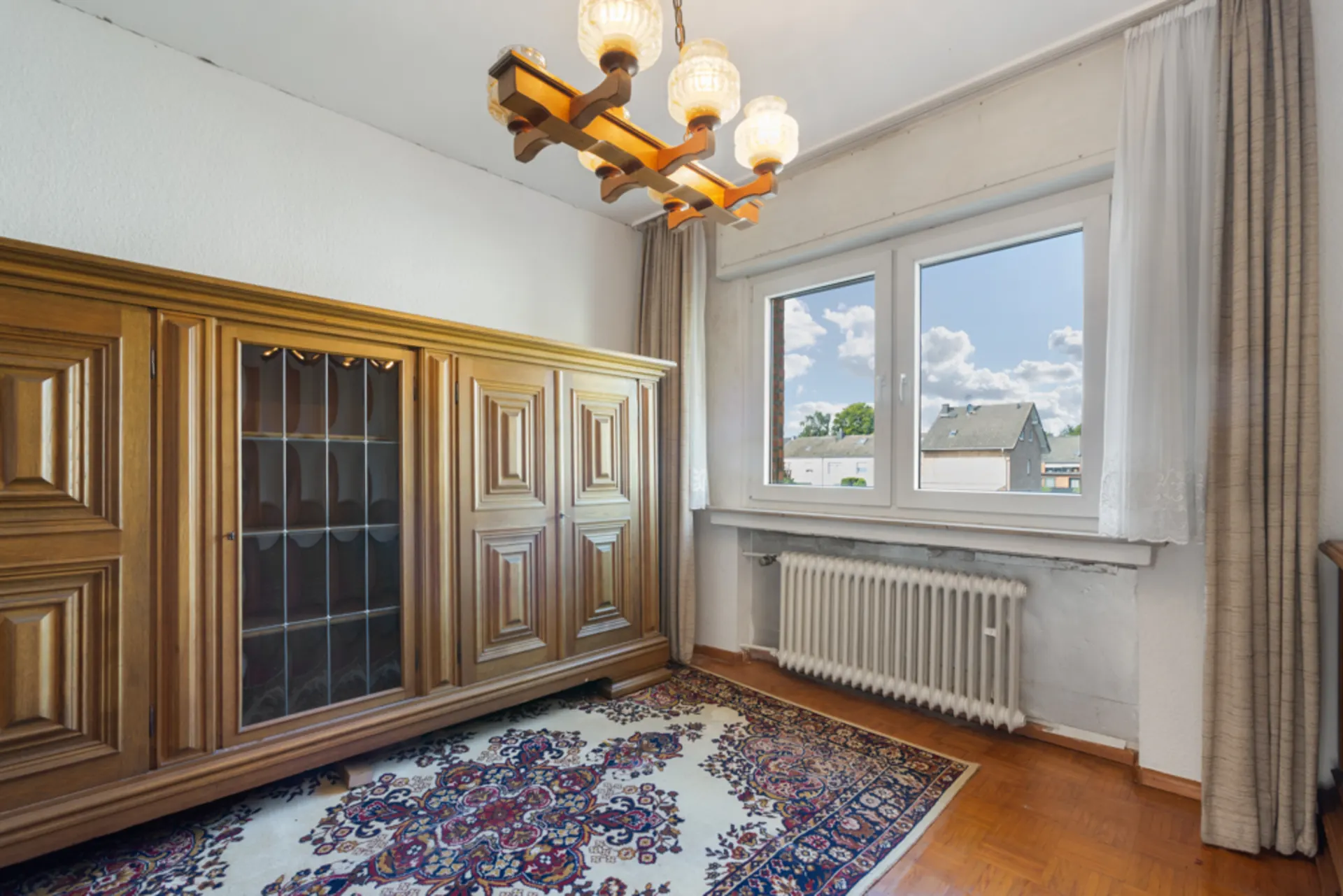 Zimmer mit Holzschrank, großem Fenster, Heizkörper und gemustertem Teppich.