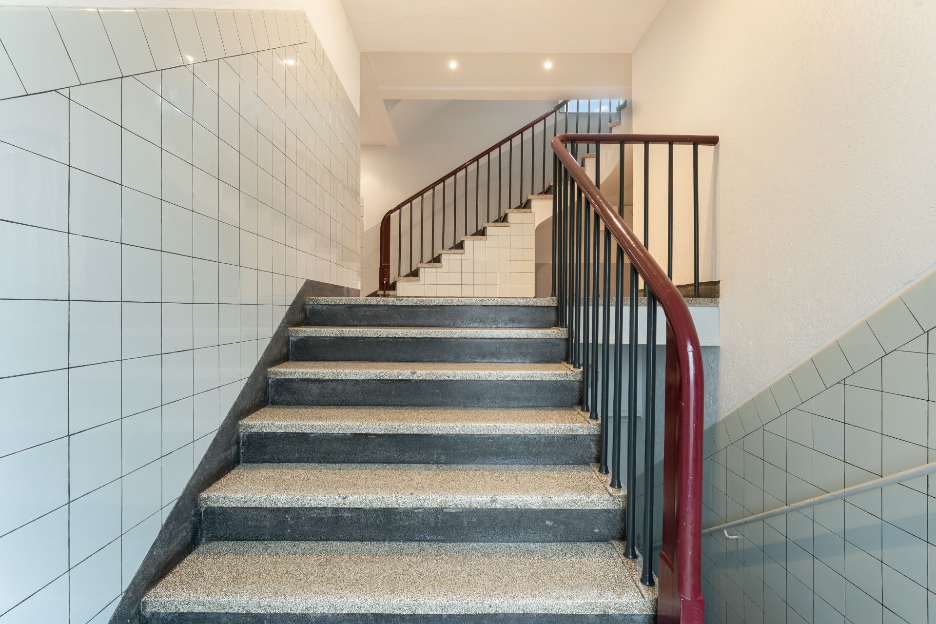 Treppenhaus mit grauen Stufen, Fliesen an den Wänden und rotem Geländer.