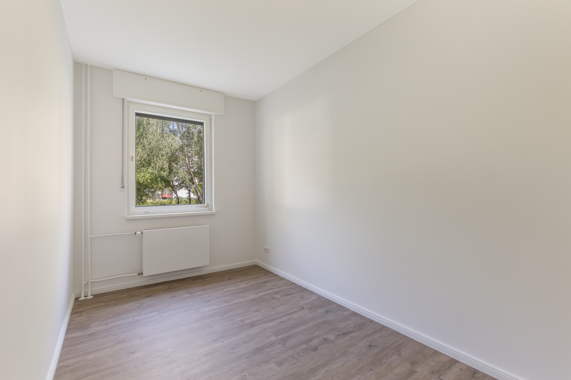 Leeres Zimmer mit hellem Boden, weißer Wand und großem Fenster mit Blick ins Grüne.