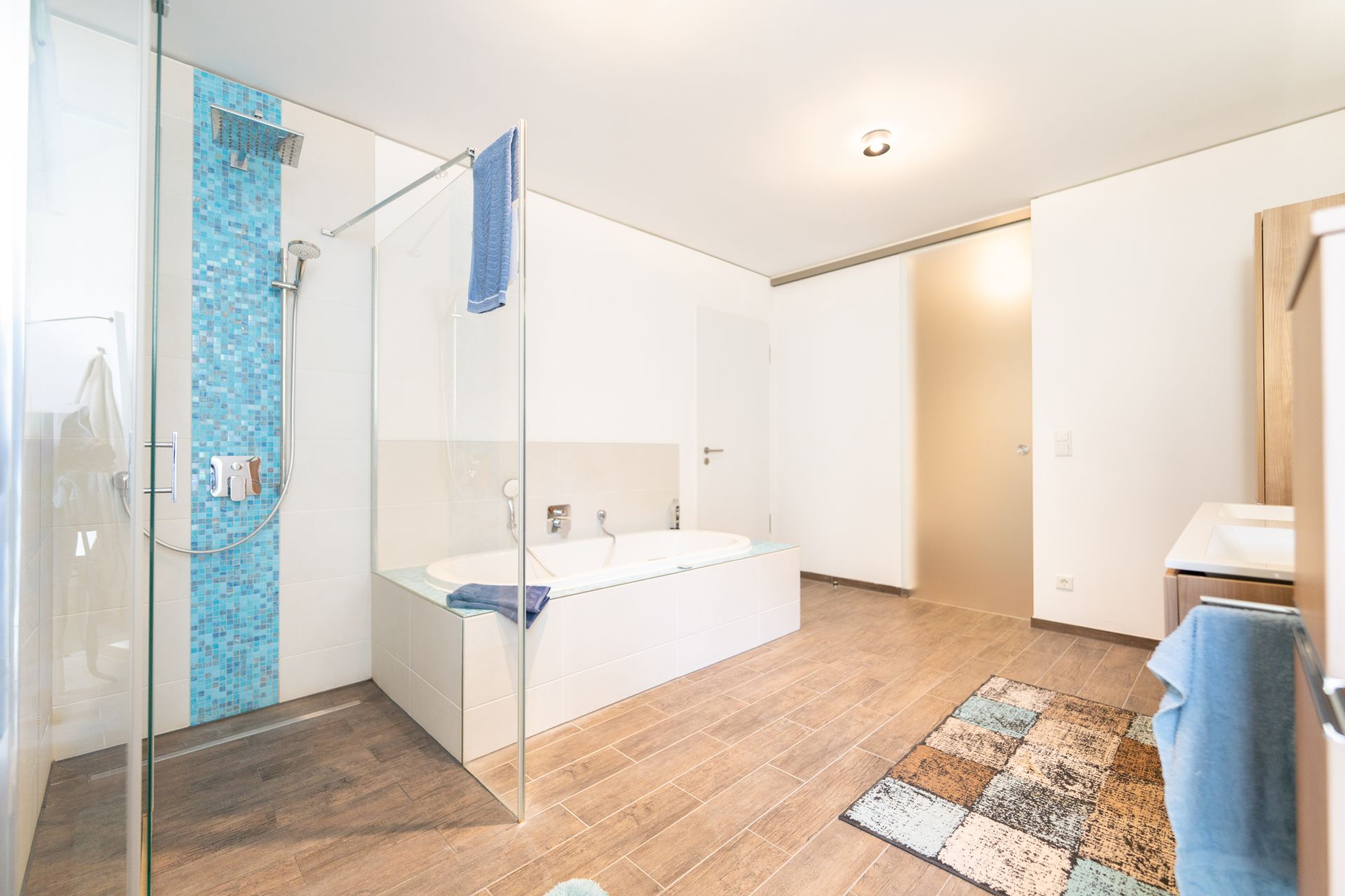 Modernes Badezimmer mit Dusche, Badewanne, Waschtisch und gemustertem Teppich auf Holzfußboden.