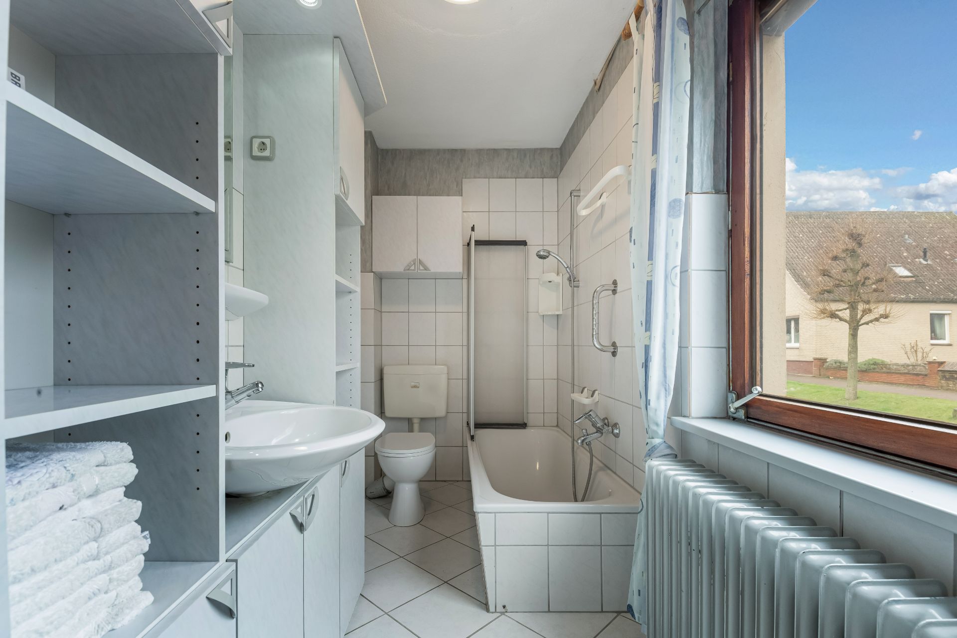 Helles Badezimmer mit Badewanne, Waschbecken, Toilette und Regal, Fenster mit Blick nach draußen.