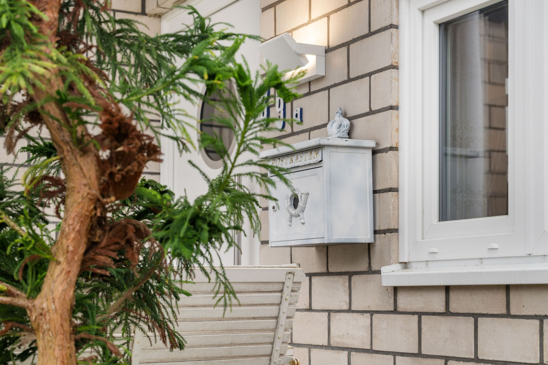 Ein Briefkasten an einer Hauswand neben einem Fenster und einer grünen Pflanze im Vordergrund.