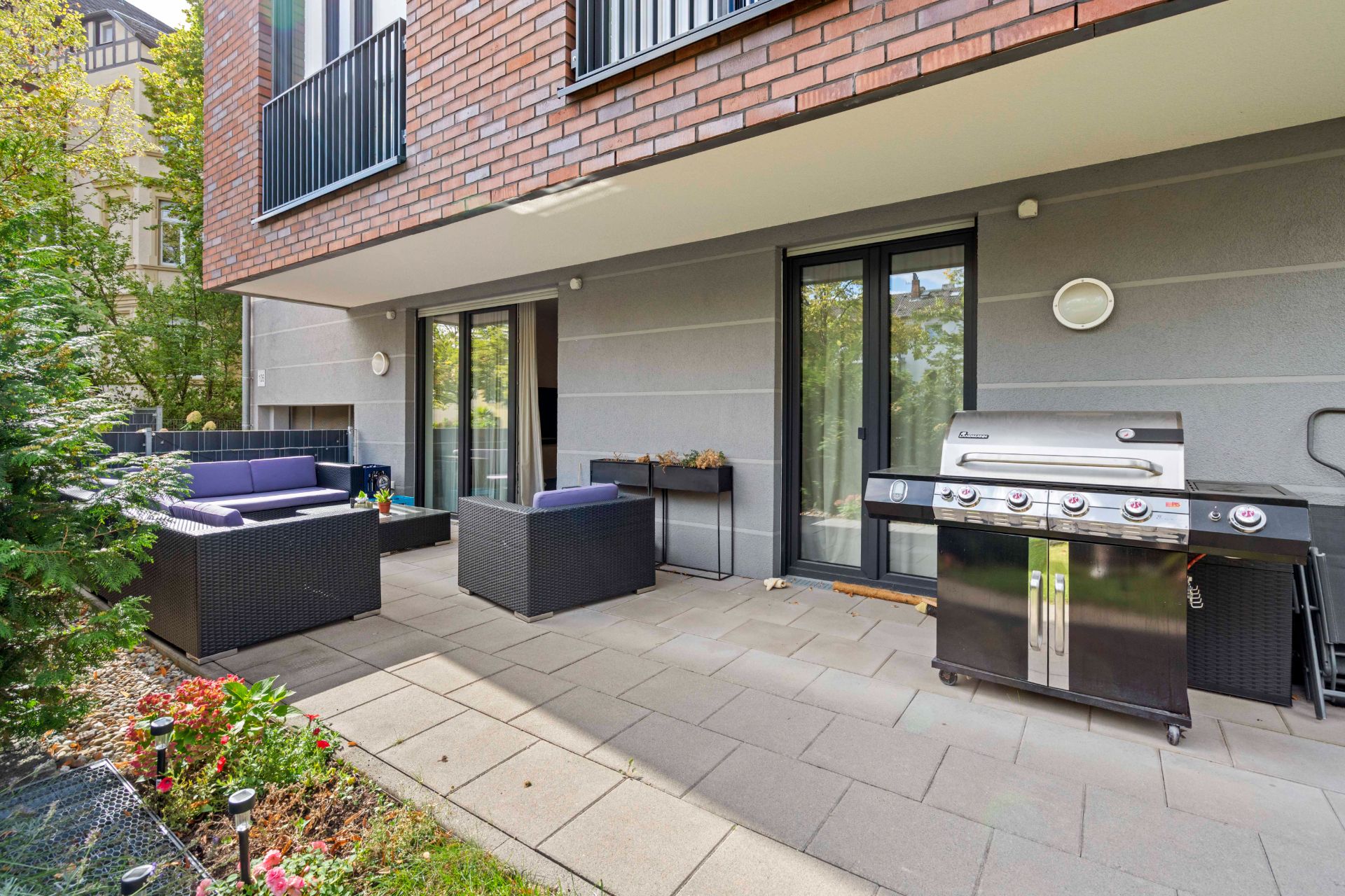 Moderne Terrasse mit Sitzmöbeln, Grill und Blumenbeet vor einer Wohnhausfassade.