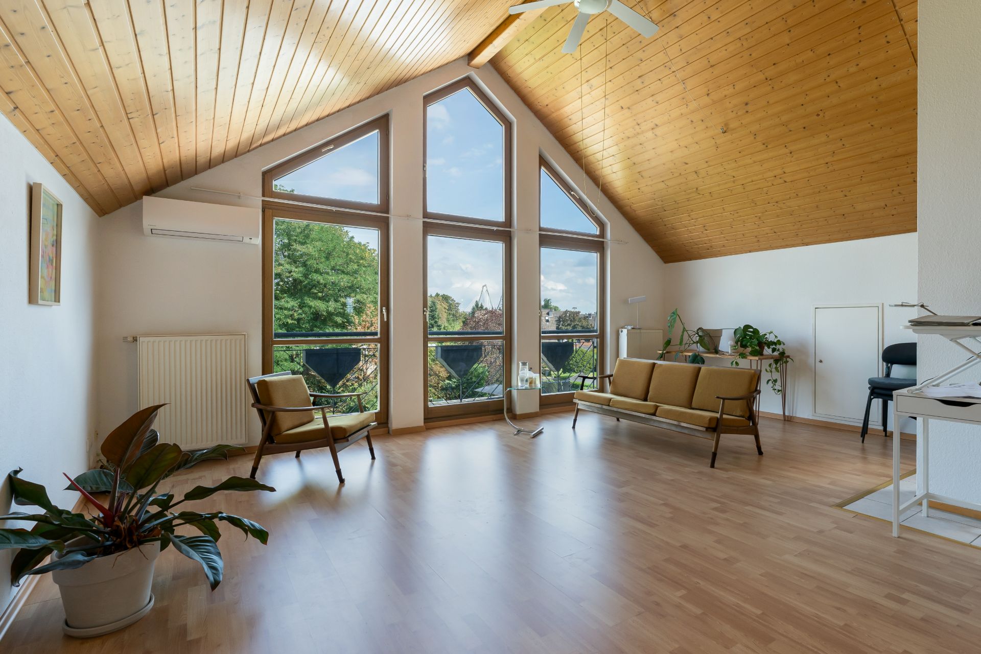 Helles Wohnzimmer mit Holzdecke, großen Fenstern, Sitzmöbeln und Zimmerpflanzen.