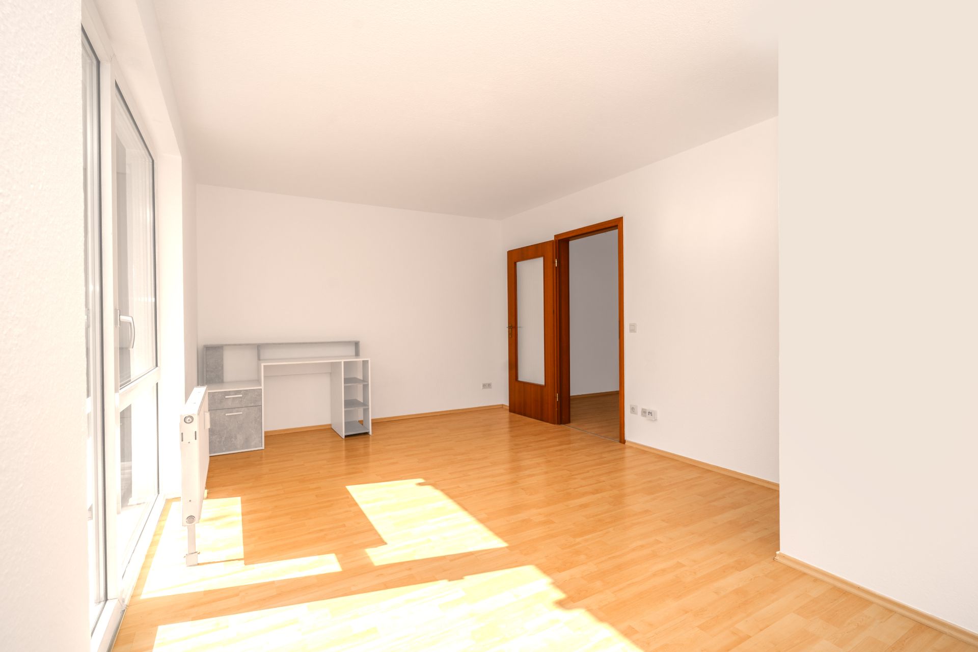 Heller Raum mit Holzboden, Schreibtisch, weißen Wänden und Glastür zu einem weiteren Zimmer.