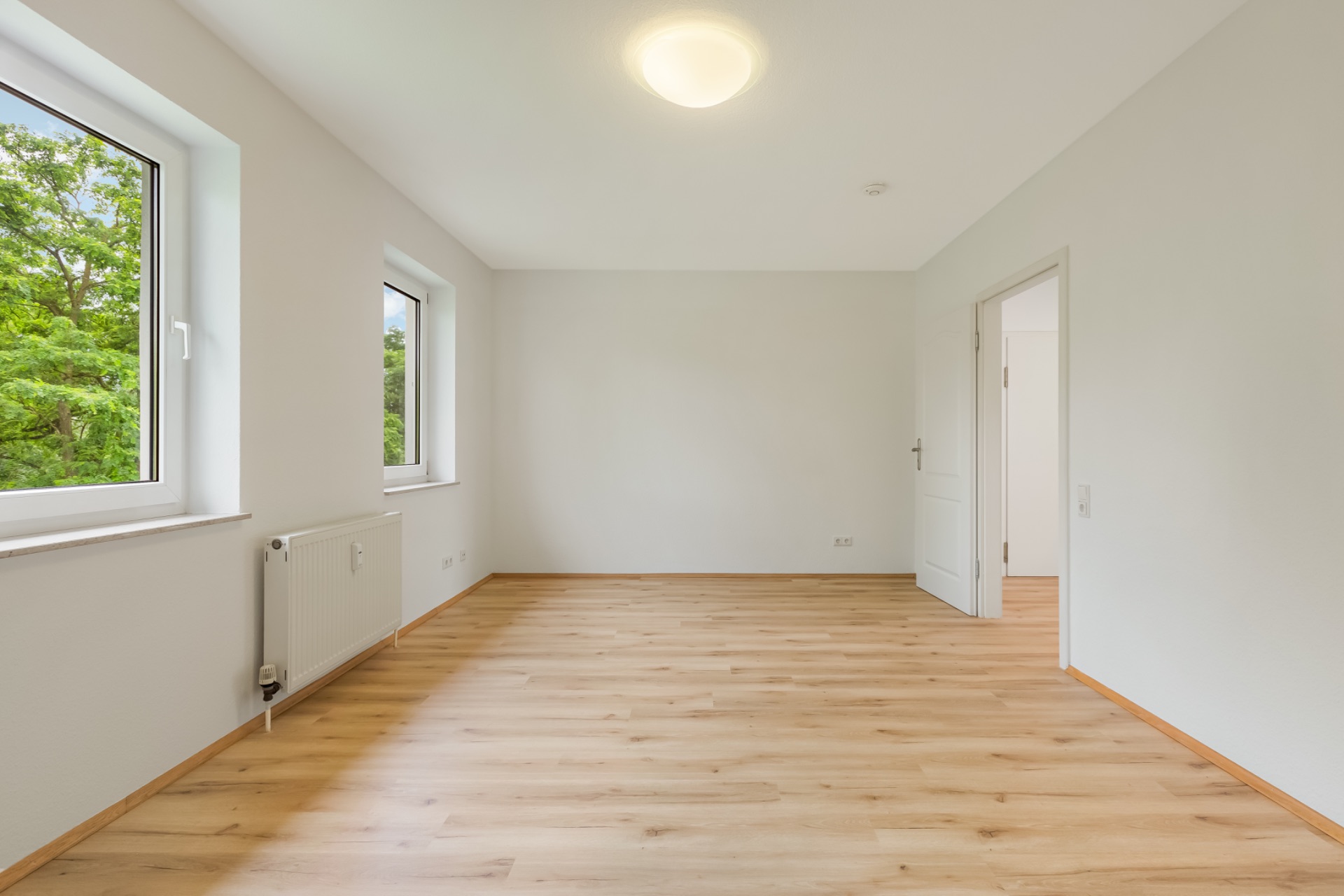 Helles, leeres Zimmer mit Holzboden, zwei Fenstern und einer weißen Tür.