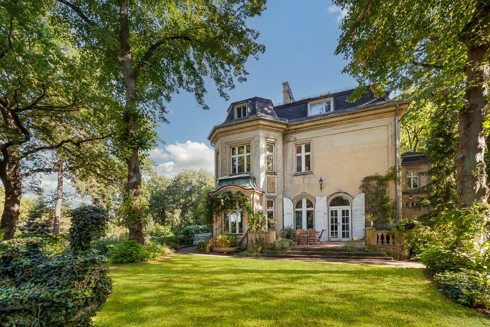 Großes Wohnhaus mit Terrasse, weißen Fenstern und Garten, umgeben von hohen Bäumen.