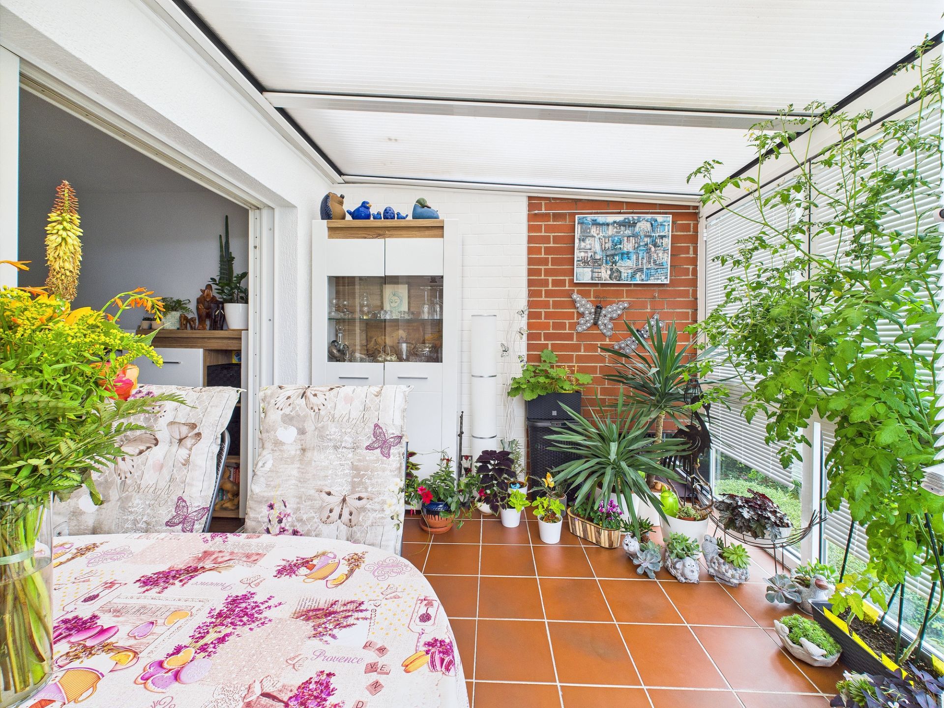 Heller Wintergarten mit vielen Zimmerpflanzen, Tisch mit Blumen und braun-rotem Fliesenboden.