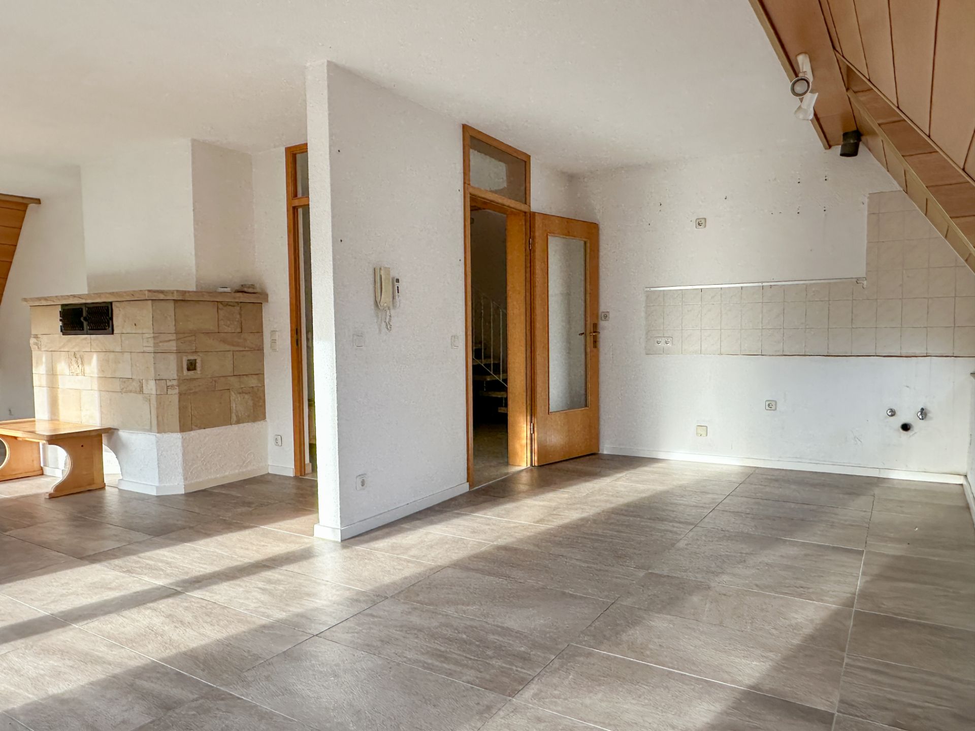 Heller Wohnraum mit Kamin, Fliesenboden, weißen Wänden und Holztür, offene Fläche zur Küche.