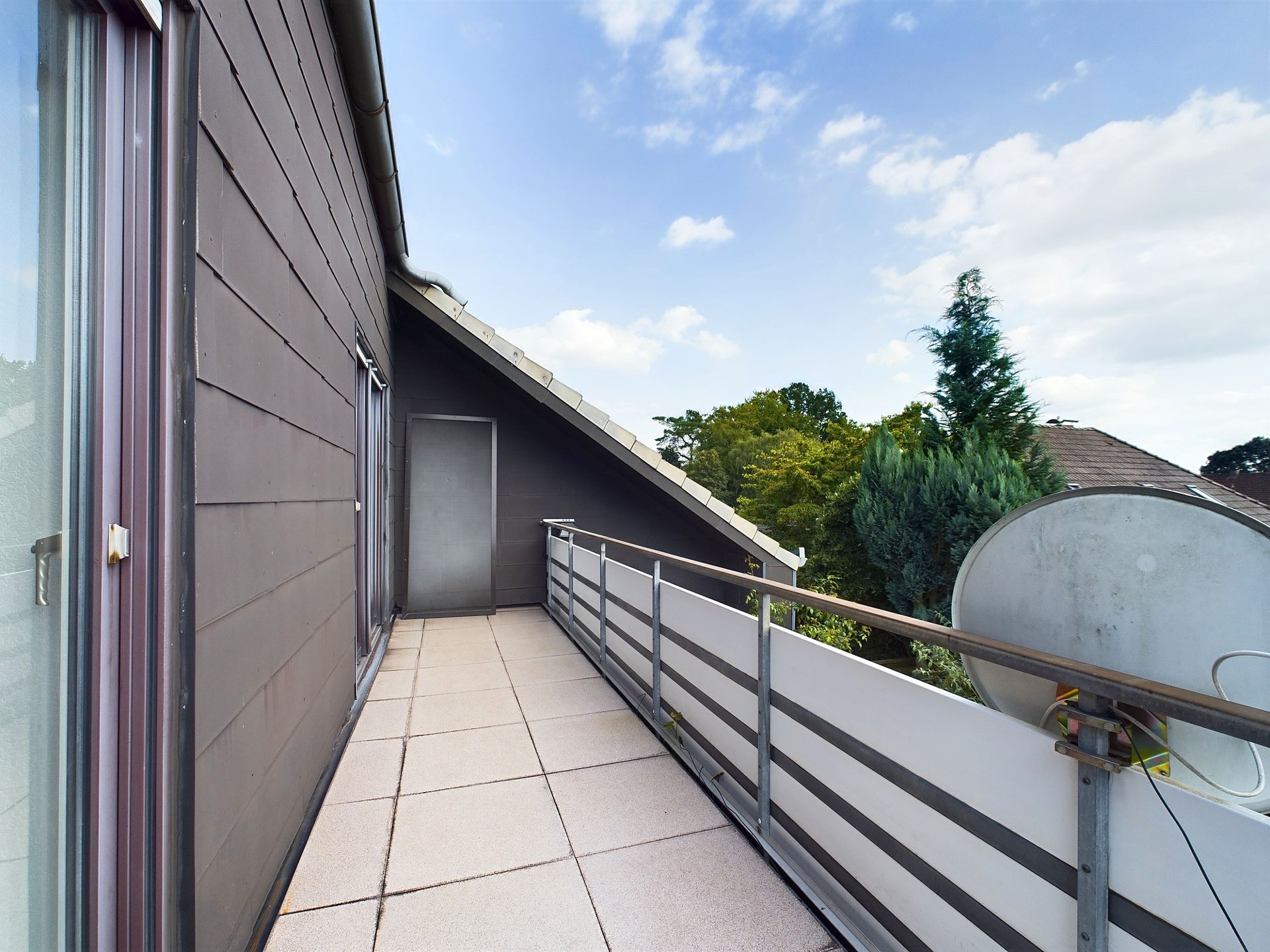 Balkon mit Fliesenboden, Metallgeländer und Satellitenschüssel, daneben Bäume und blauer Himmel.