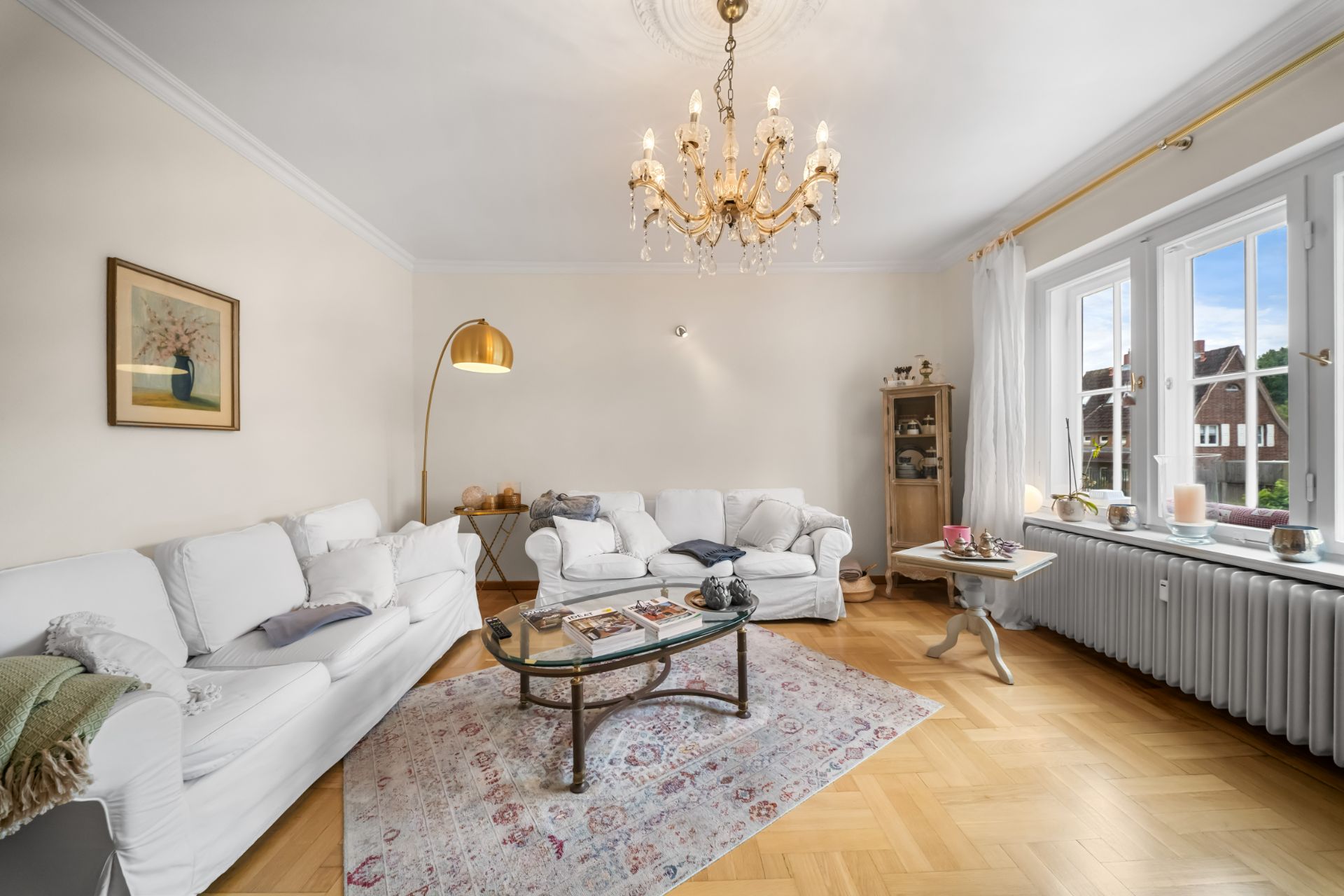 Helles Wohnzimmer mit weißen Sofas, Glastisch, Teppich, Kronleuchter und großem Fenster.