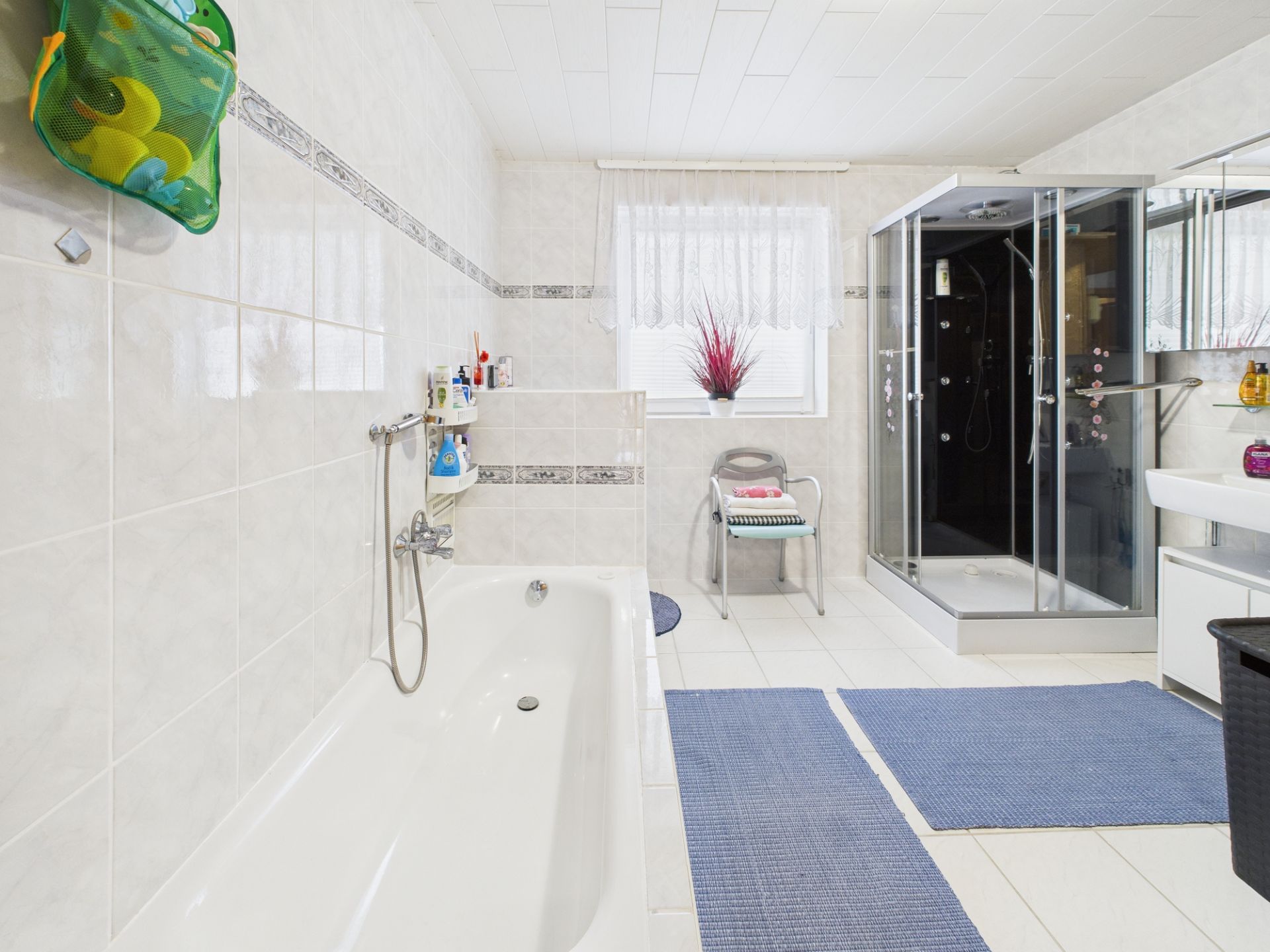 Helles Badezimmer mit Badewanne, Dusche, Waschbecken, Fenster und blauen Teppichen.