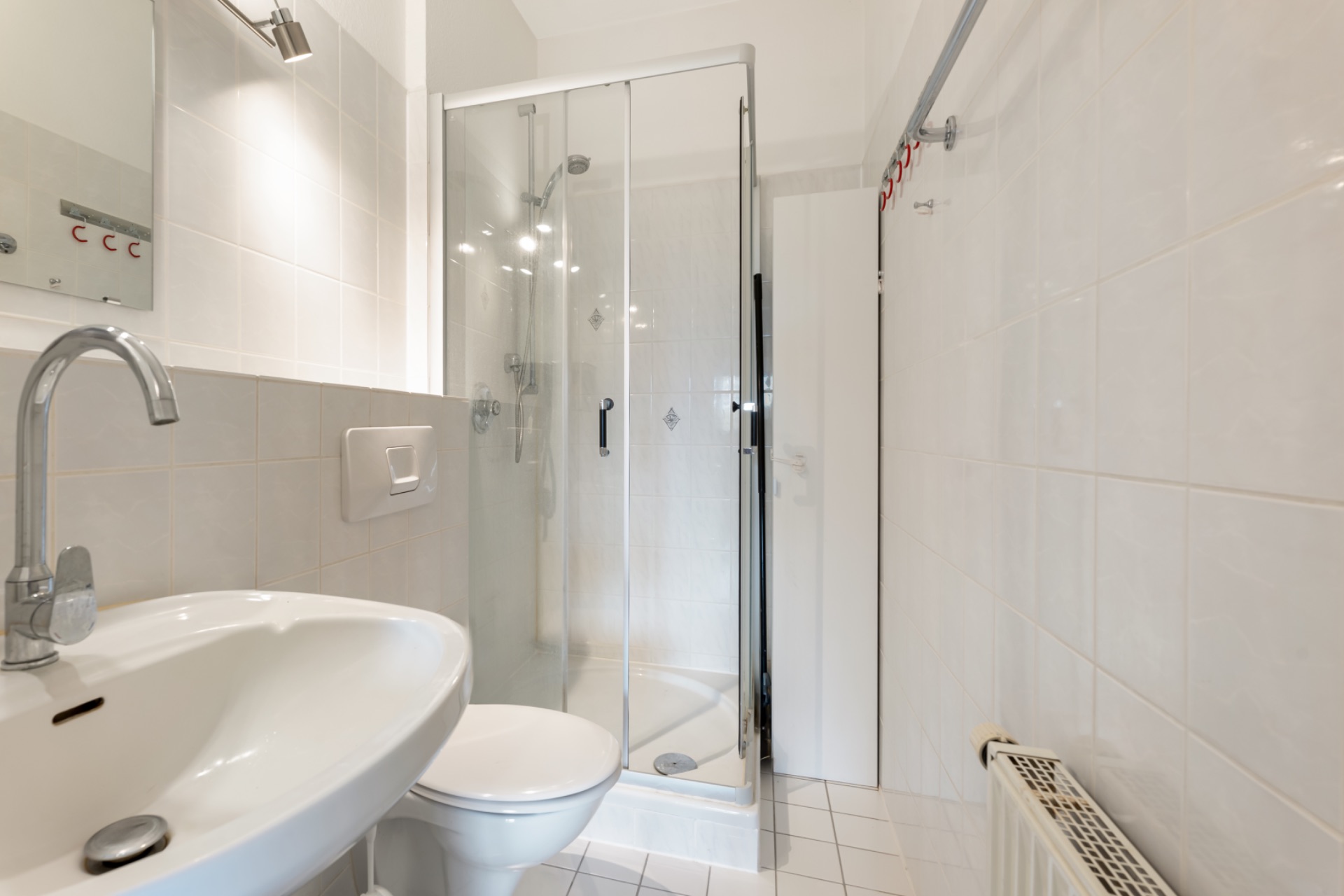 Helles Badezimmer mit Waschbecken, WC, Dusche mit Glastür und Heizkörper.