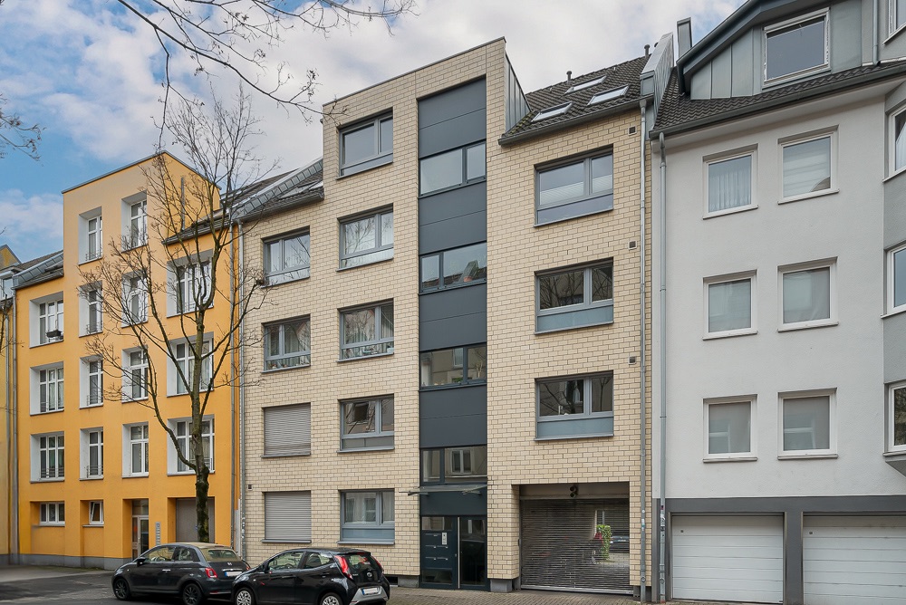 Mehrstöckiges Wohnhaus mit heller Fassade, großen Fenstern und geparkten Autos davor.