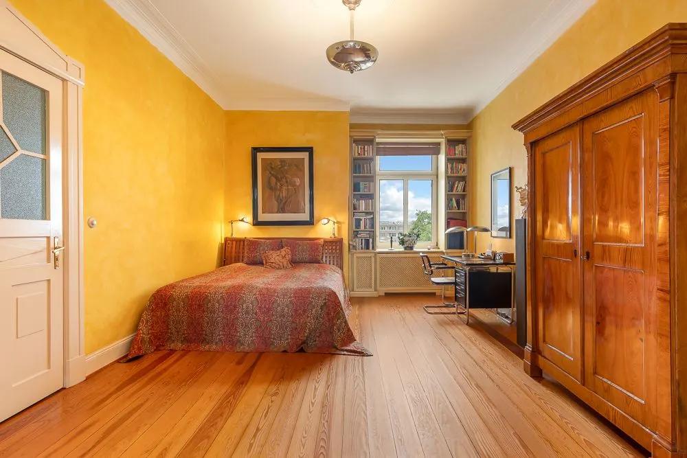 Helles Schlafzimmer mit Einzelbett, Holzboden, großem Schrank und Schreibtisch vor dem Fenster.