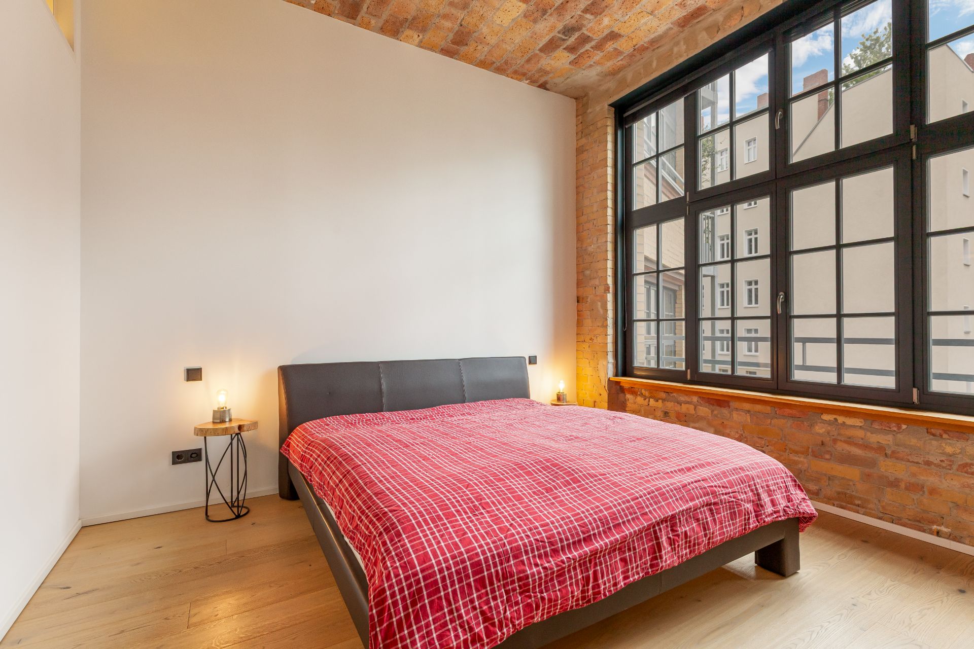 Schlafzimmer mit großem Fenster, Bett mit roter Decke und Nachttisch mit Lampe.