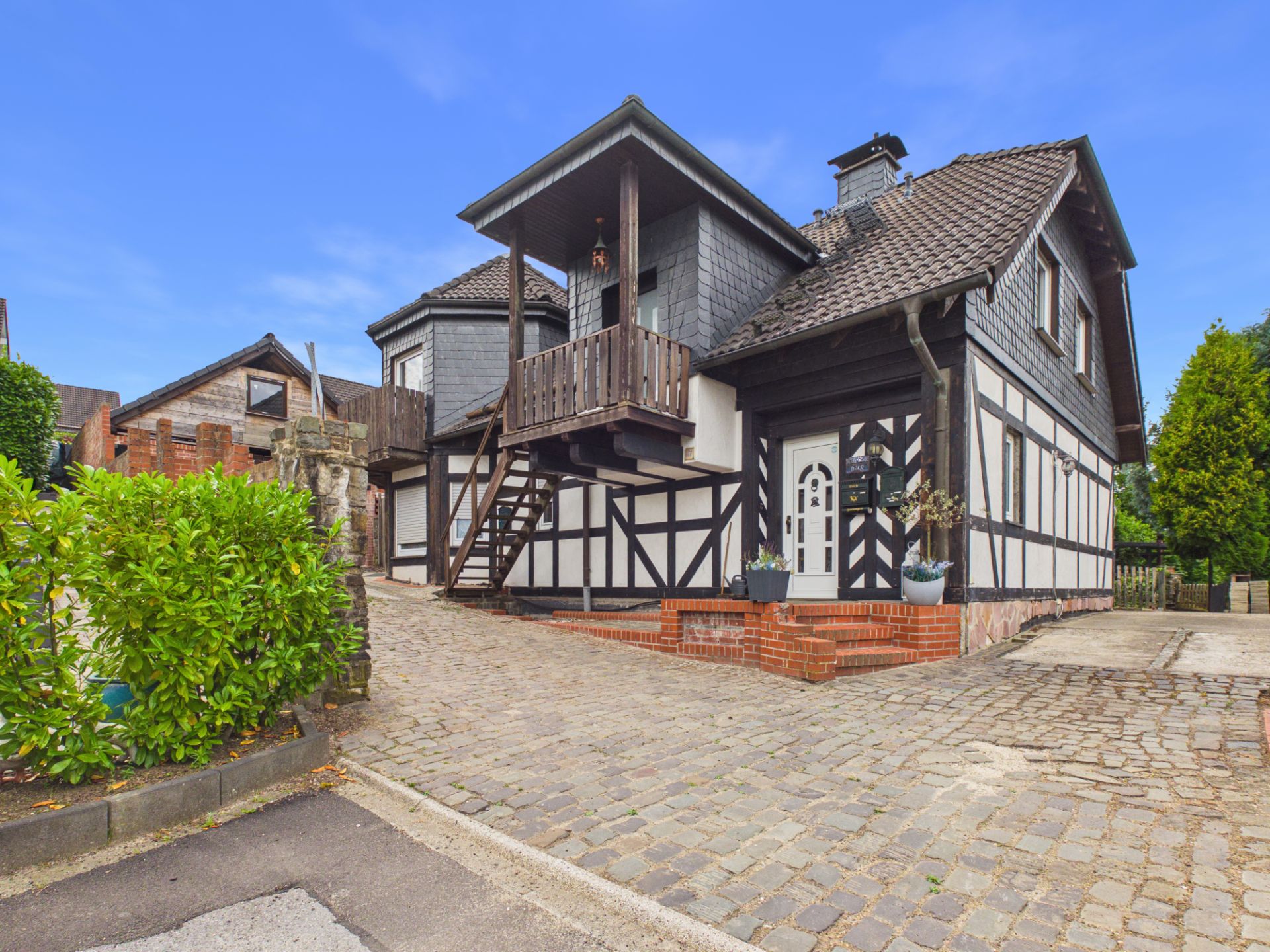 Fachwerkhaus mit dunklem Dach, Eingangsbereich und kleinem Balkon, gepflasterte Einfahrt davor.