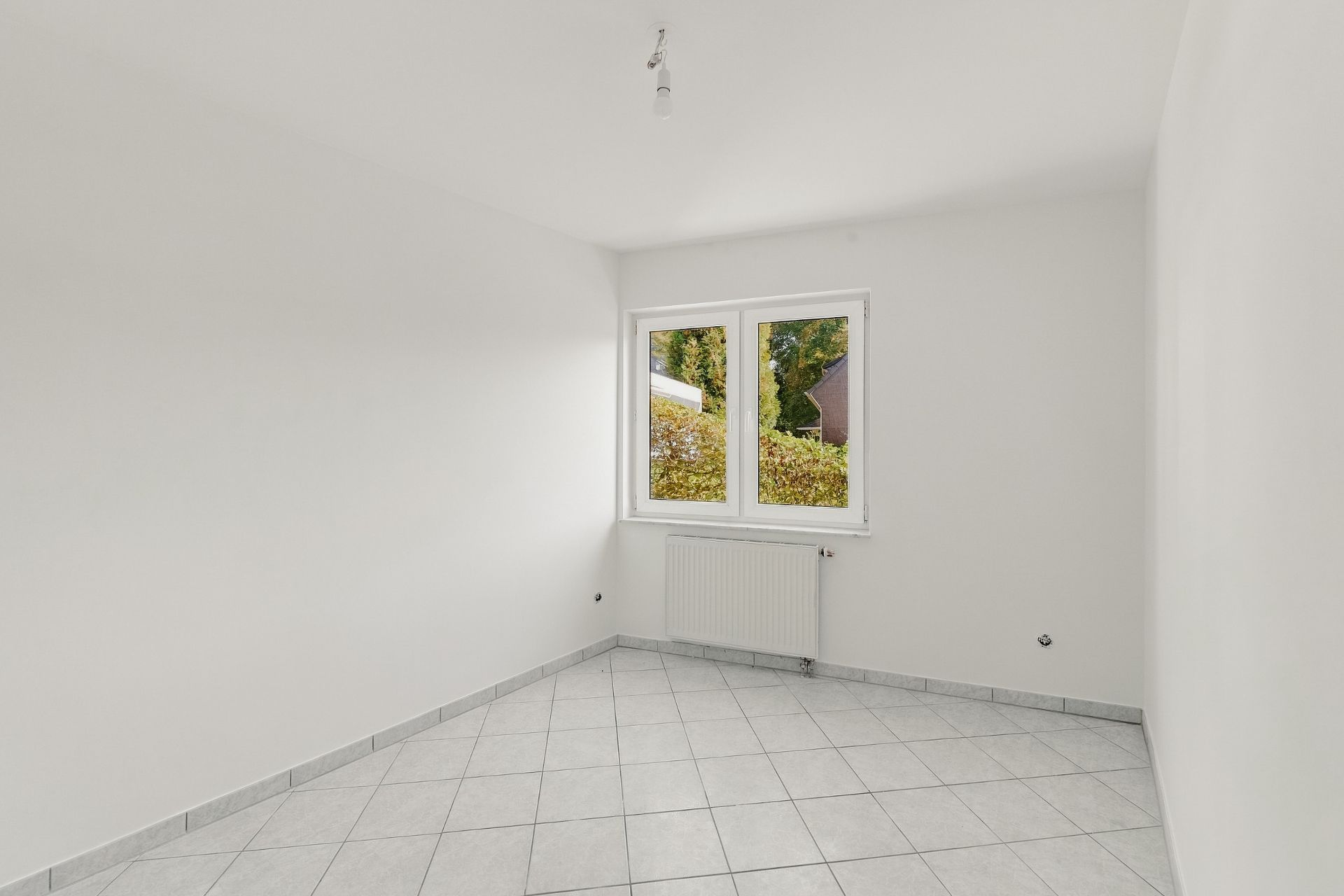 Heller, leerer Raum mit weißen Fliesen, weißer Wand und Fenster mit Blick ins Grüne.
