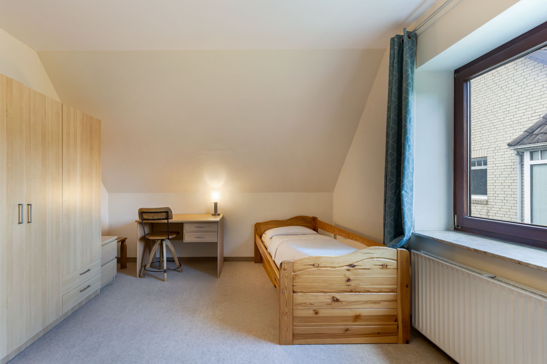 Helles Schlafzimmer mit Holzbett, Schreibtisch, Stuhl, Kleiderschrank und großem Fenster mit Vorhang.