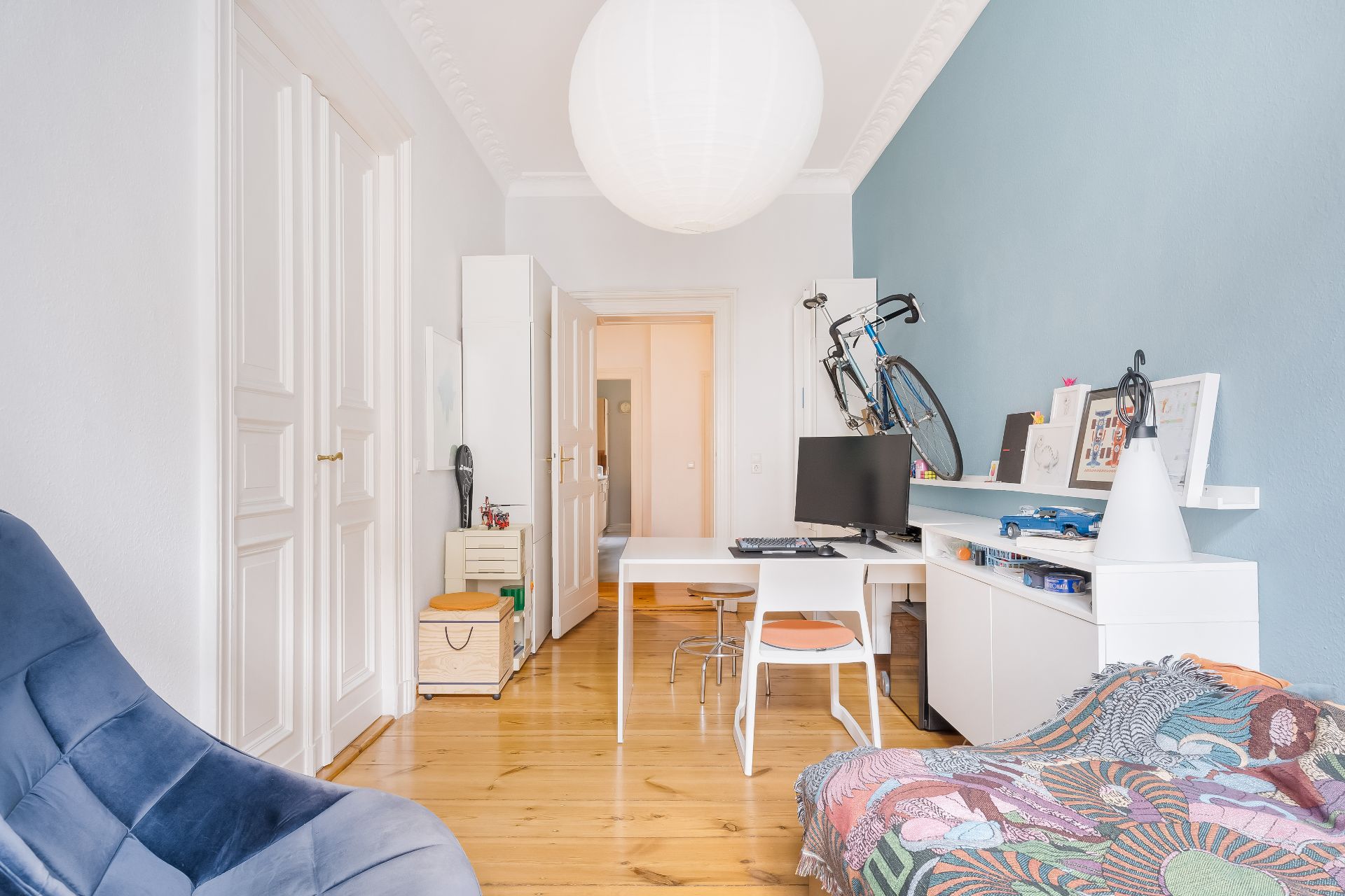 Helles Zimmer mit Schreibtisch, Computer, Fahrrad an der Wand und bunter Decke auf dem Bett.