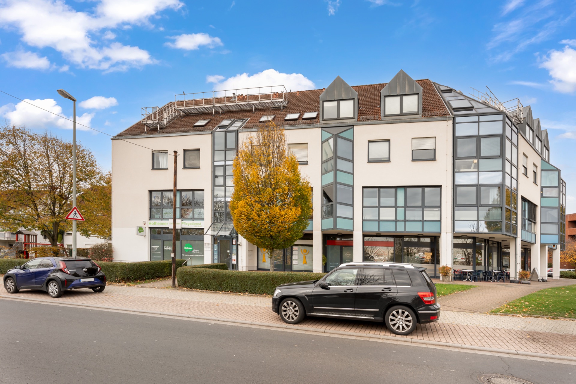 Modernes Wohngebäude mit großen Fenstern, Bäumen davor und geparkten Autos an der Straße.