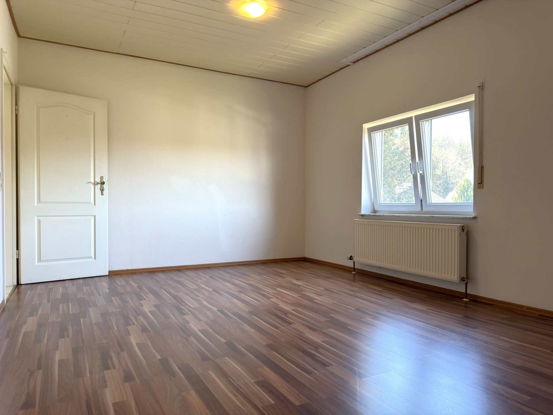 Helles, leeres Zimmer mit Holzfußboden, weißer Tür und Fenster mit Heizkörper.