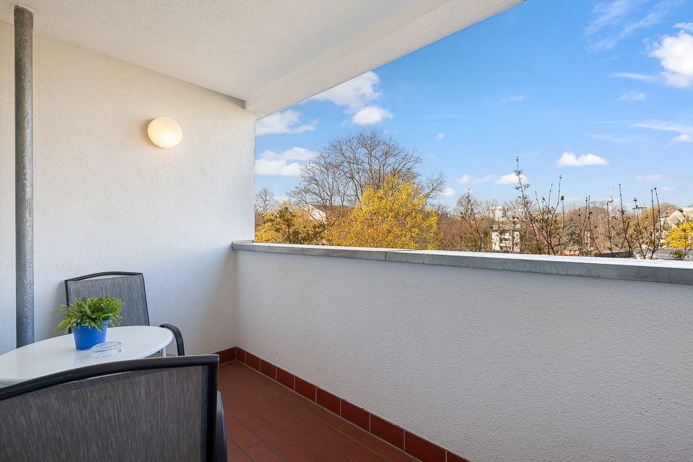 Balkon mit Tisch, zwei Stühlen und einer Pflanze, Ausblick auf Bäume und blauen Himmel.