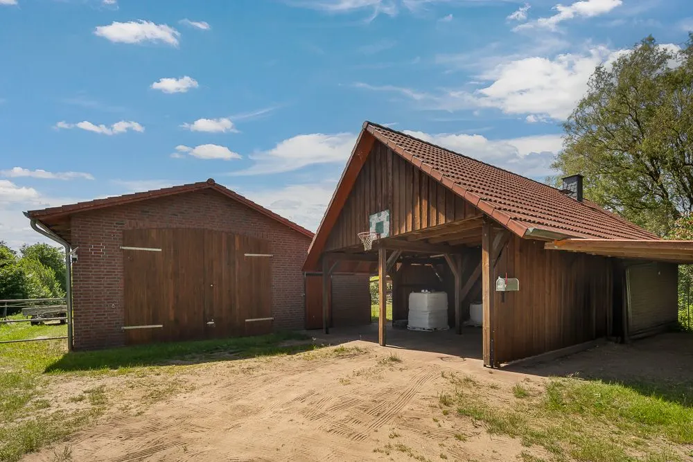Holzunterstand mit Satteldach neben rotem Backsteingebäude, Sandboden im Vordergrund.
