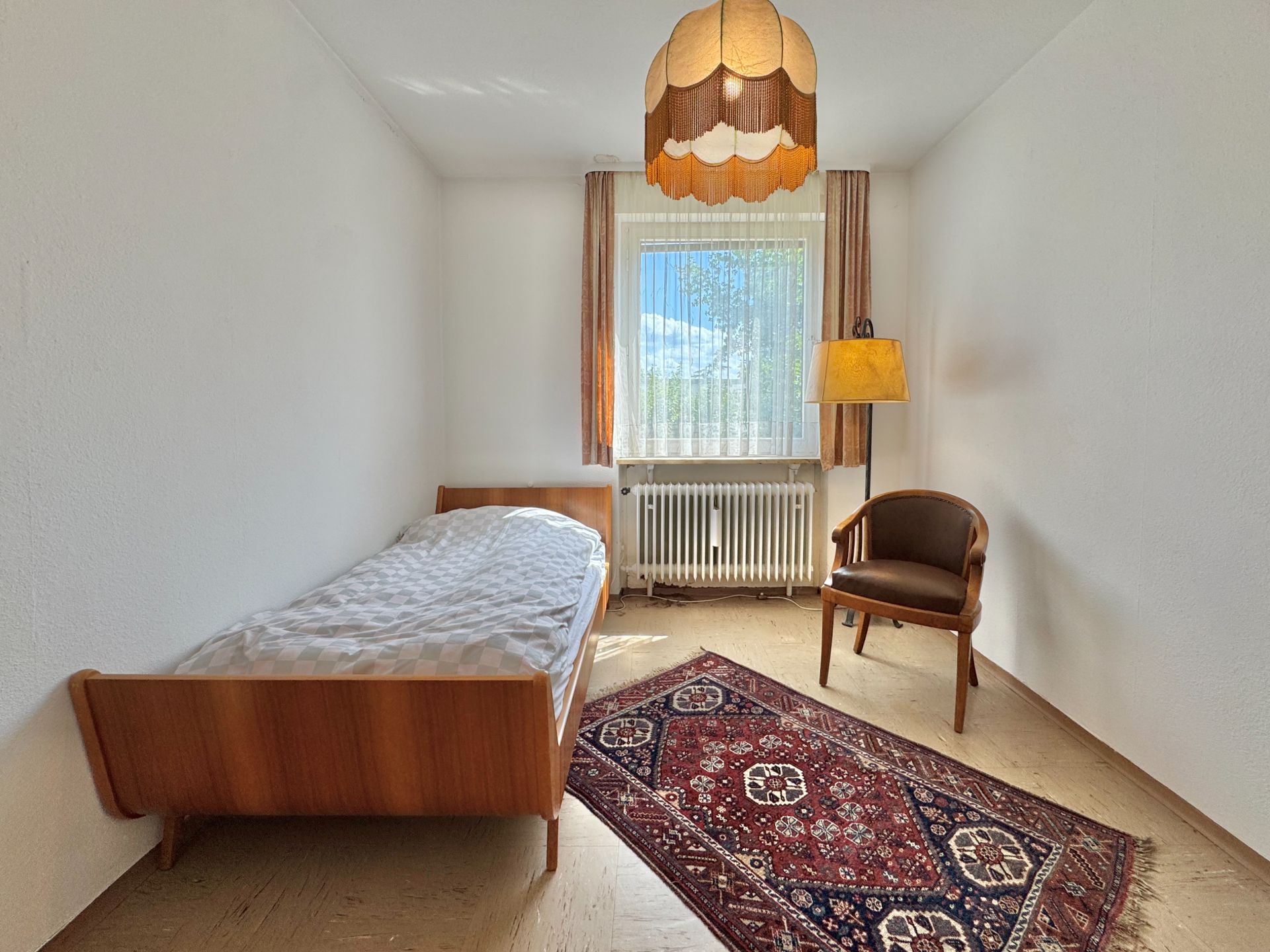 Helles Schlafzimmer mit Einzelbett, Teppich, Stuhl und Fenster mit Vorhängen.