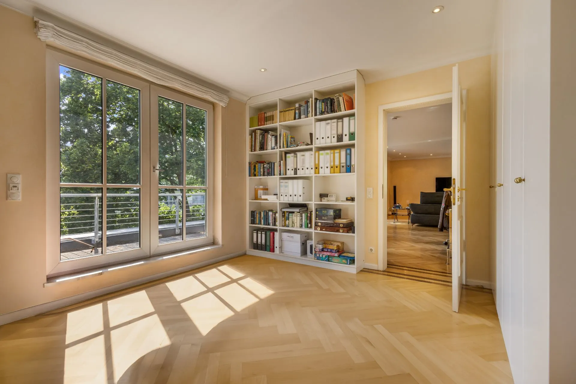 Helles Zimmer mit Bücherregal, Parkettboden und großen Fenstern mit Blick auf einen Balkon.