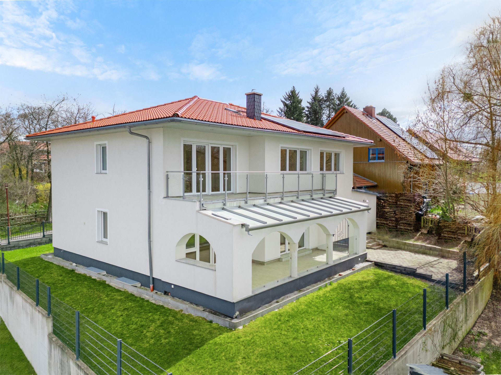 Modernes zweistöckiges weißes Haus mit rotem Dach, Balkon und überdachter Terrasse im Garten.