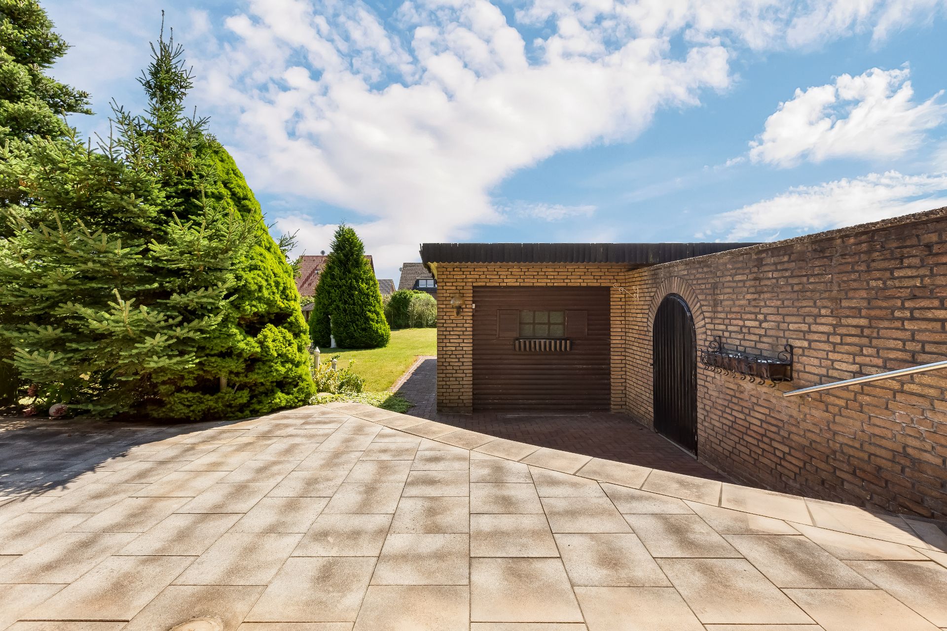 Gepflasterte Terrasse mit gemauerter Garage, Tür und Garten mit Tannenbäumen im Hintergrund.