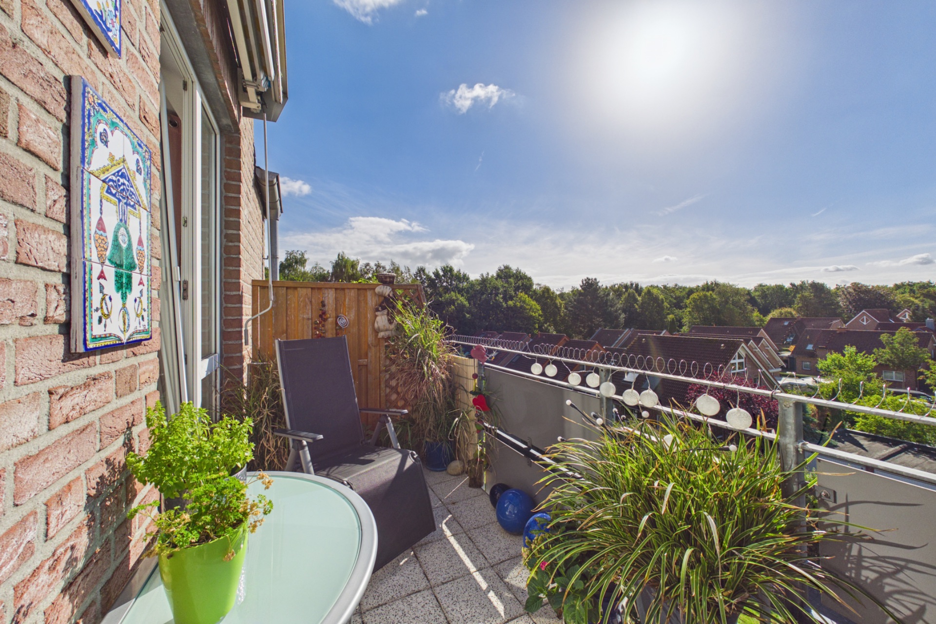 Sonniger Balkon mit Pflanzen, Stuhl und kleinem Tisch, Blick auf Dächer und Bäume.
