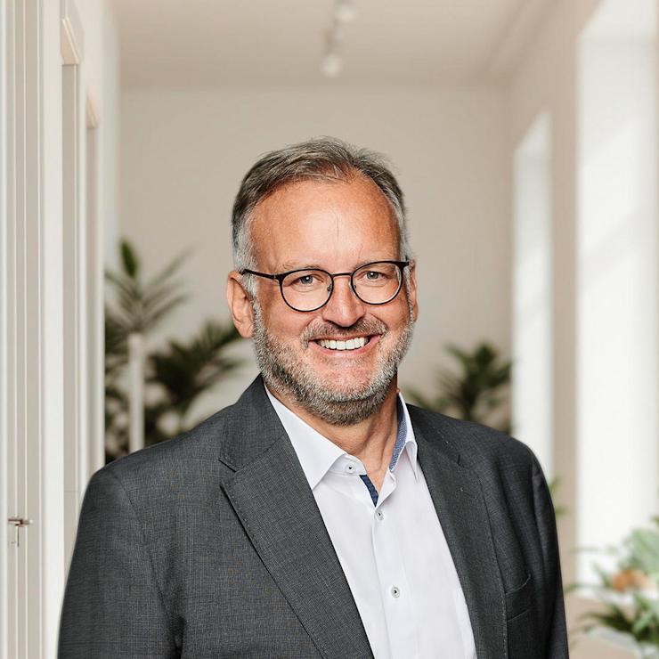 Mann mit Brille und Anzug steht in hellem Wohnraum mit Pflanzen im Hintergrund.