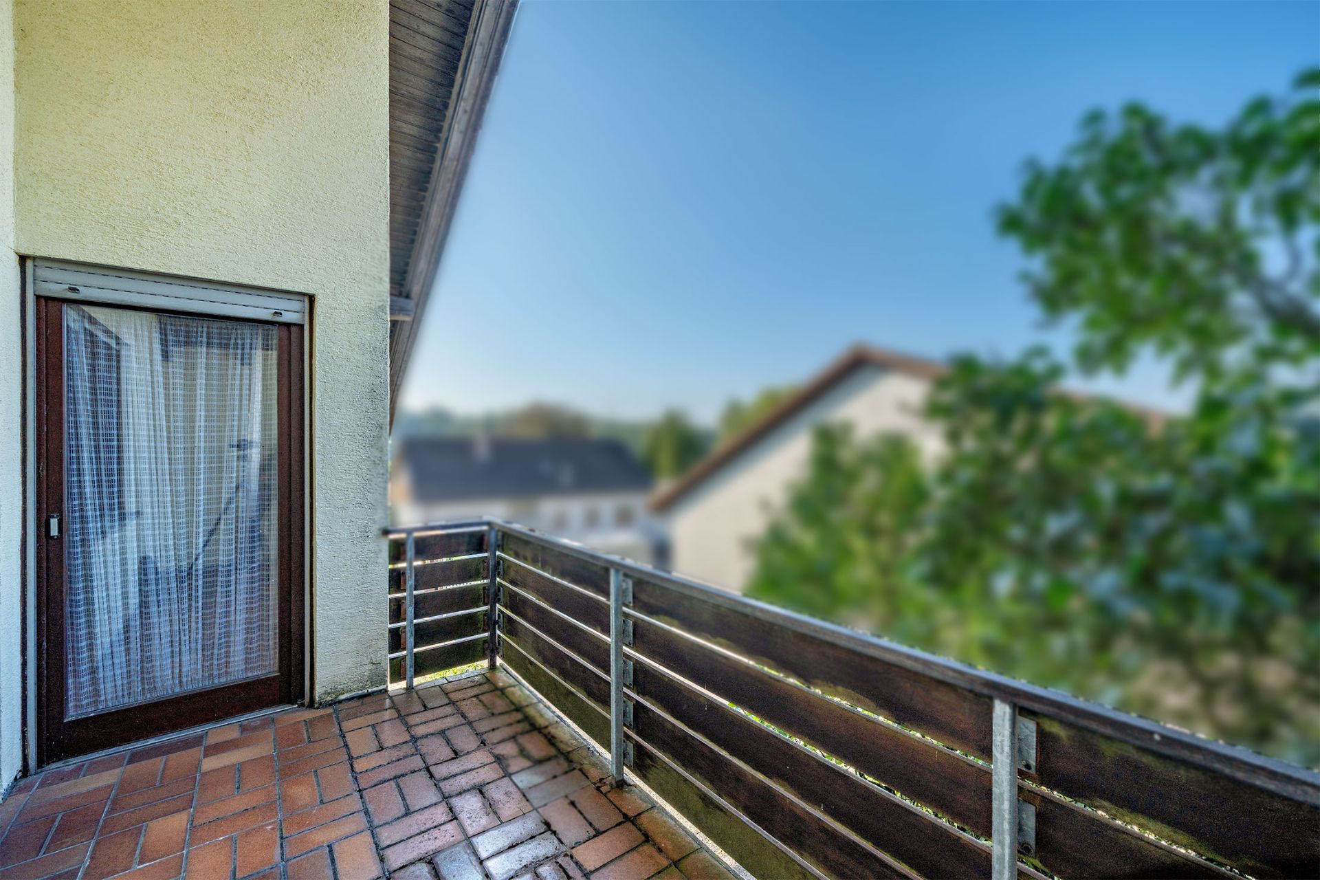 Balkon mit braunen Fliesen, Metallgeländer und Tür, daneben Bäume und Nachbarhaus sichtbar.