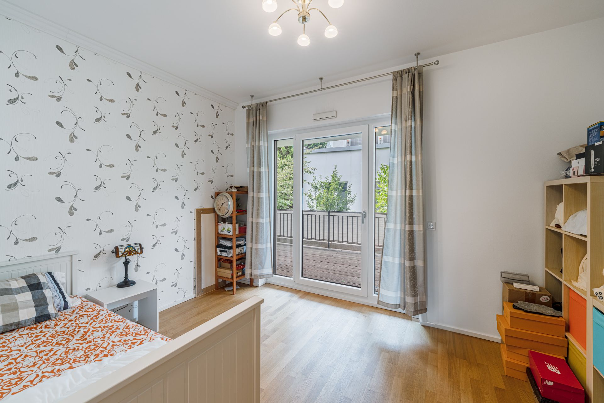 Helles Schlafzimmer mit Holzboden, Bett, Regal und Zugang zu einem Balkon durch große Glastüren.
