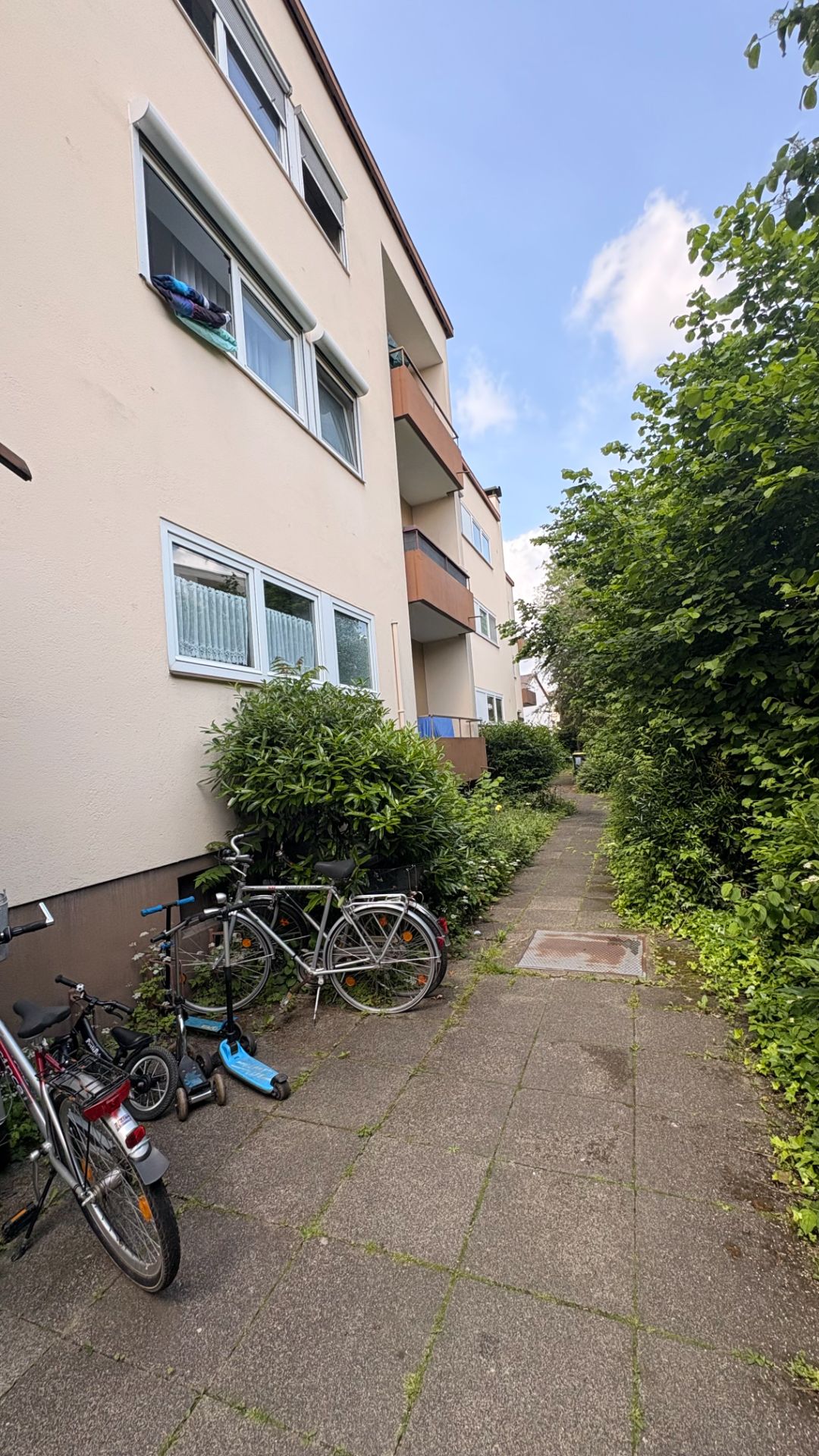 Mehrfamilienhaus mit Balkonen, Gehweg und Fahrrädern an der Hauswand, umgeben von Büschen.