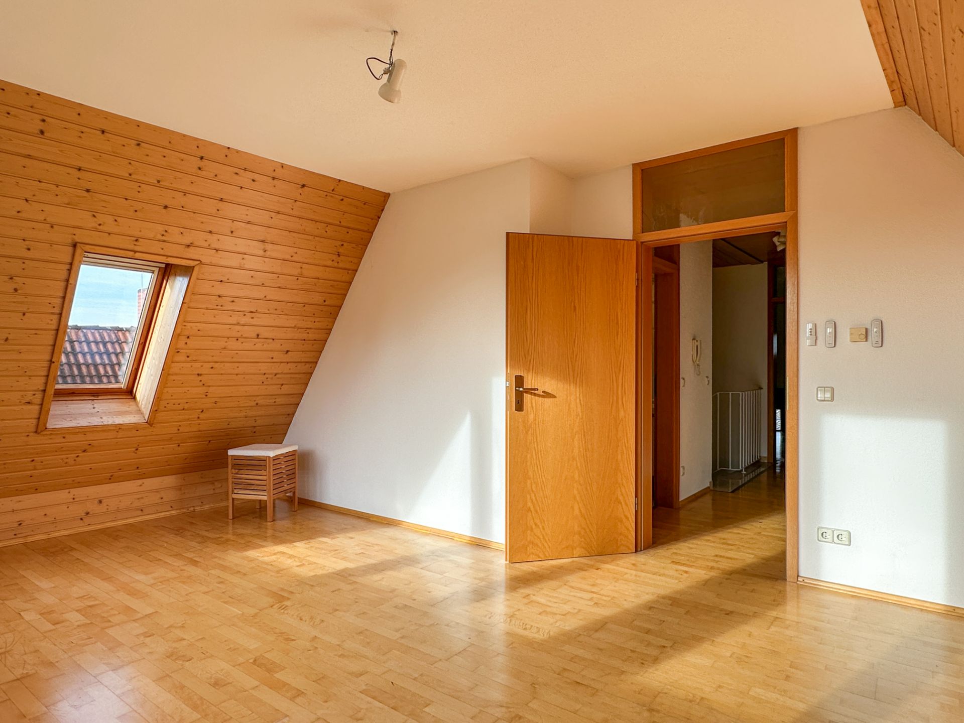 Helles Dachzimmer mit schräger Holzdecke, Dachfenster, Holztür und kleinem Hocker.