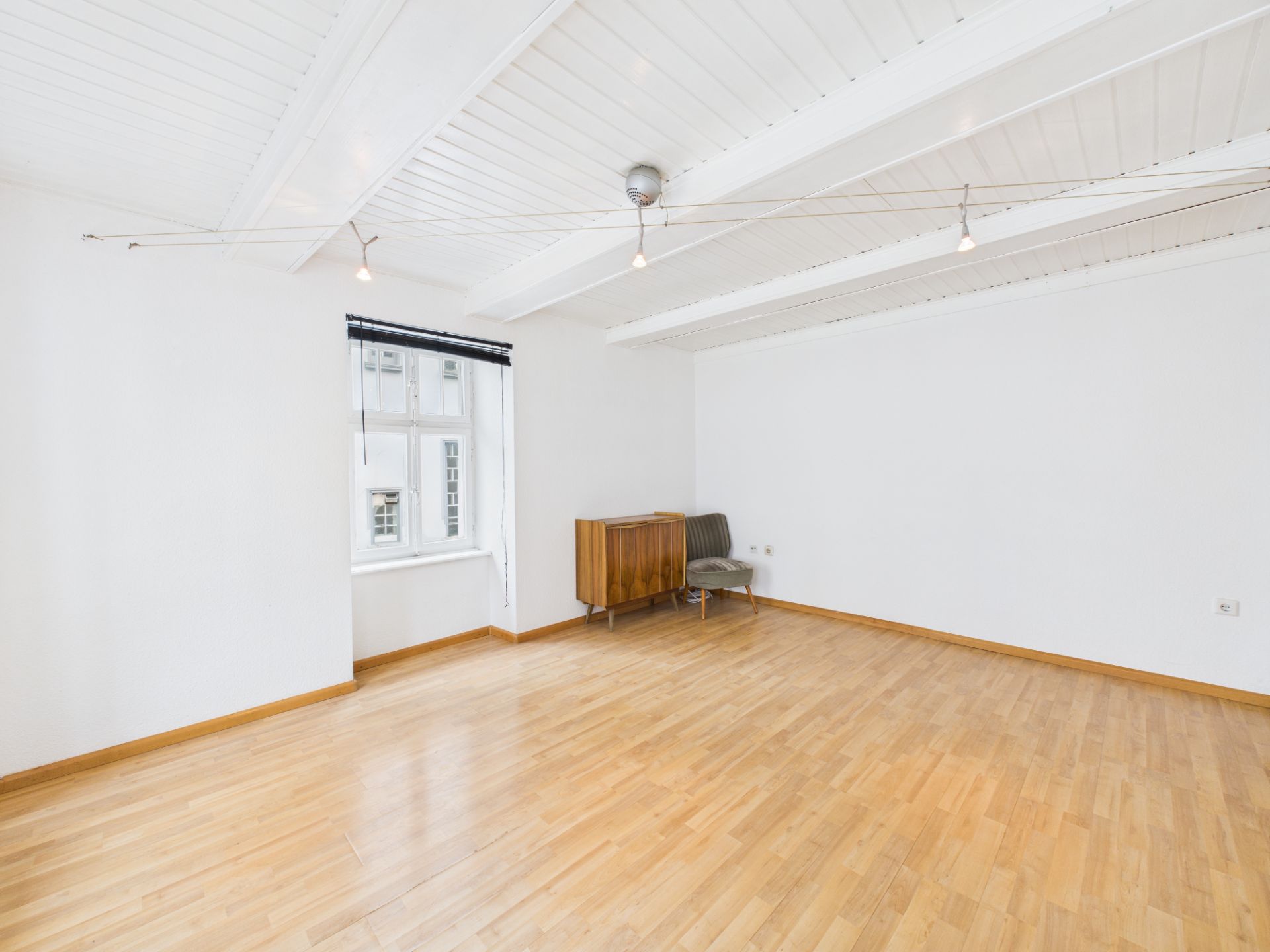 Heller Raum mit Holzfußboden, weißen Wänden, Deckenbalken, Fenster, Stuhl und kleinem Schrank.