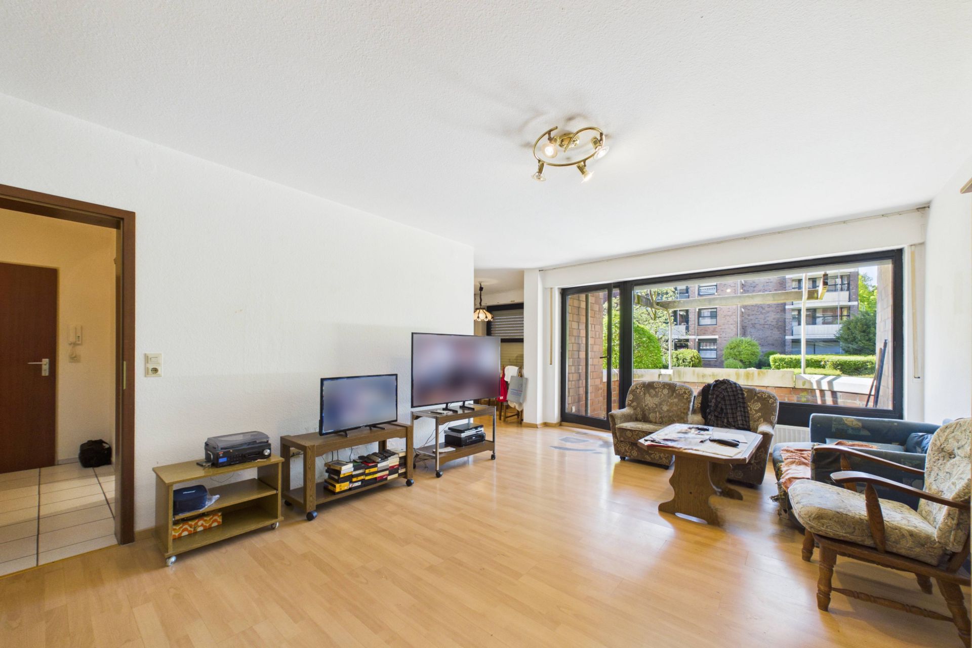 Helles Wohnzimmer mit Parkettboden, zwei Fernsehern, Sitzmöbeln und großem Fenster zur Terrasse.