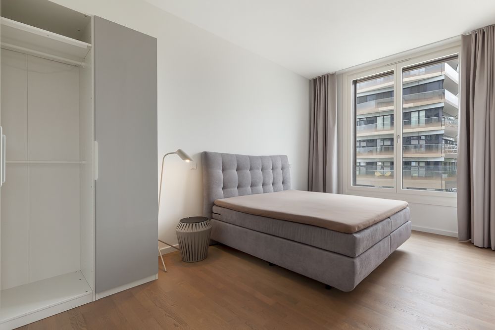 Helles Schlafzimmer mit grauem Bett, offenem Kleiderschrank und großem Fenster mit Vorhängen.