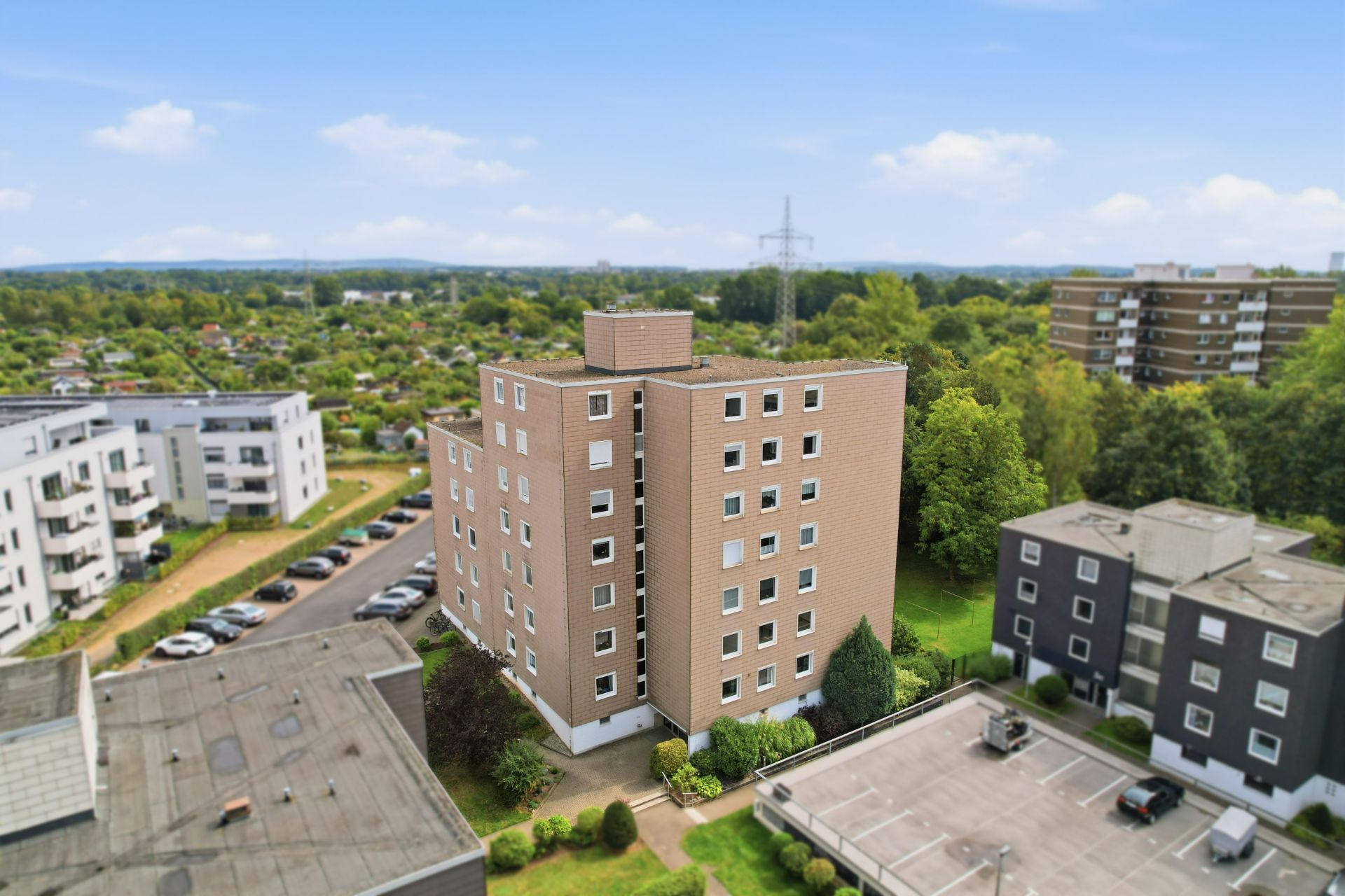 Mehrstöckiges Wohngebäude mit brauner Fassade, umgeben von Bäumen, Parkplätzen und weiteren Wohnhäusern.