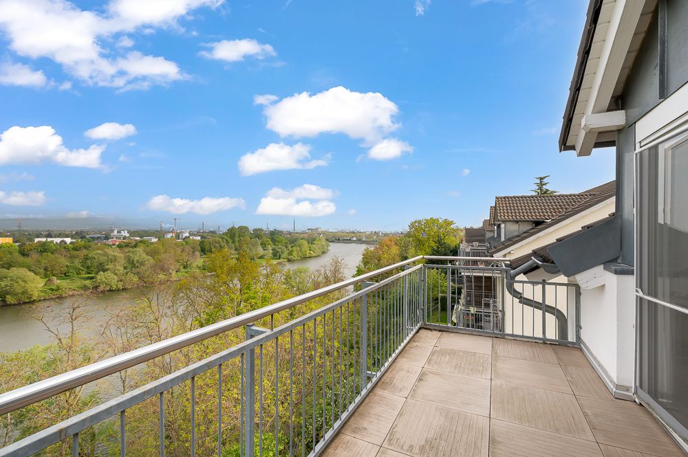 Balkon mit Geländer und Flussblick, umgeben von Bäumen, unter blauem Himmel.