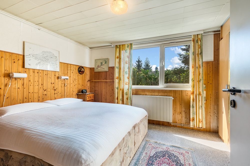 Helles Schlafzimmer mit Doppelbett, Holzwand, großem Fenster und Teppichboden.