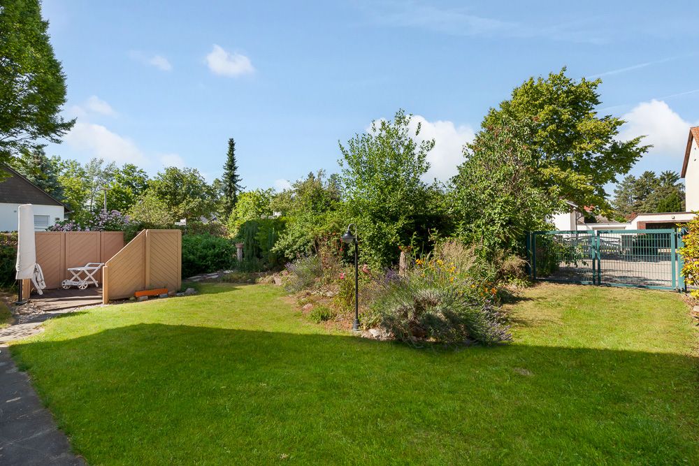 Grüner Garten mit Rasenfläche, Blumenbeet, Bäumen und einer kleinen Terrasse mit Sitzmöbeln.