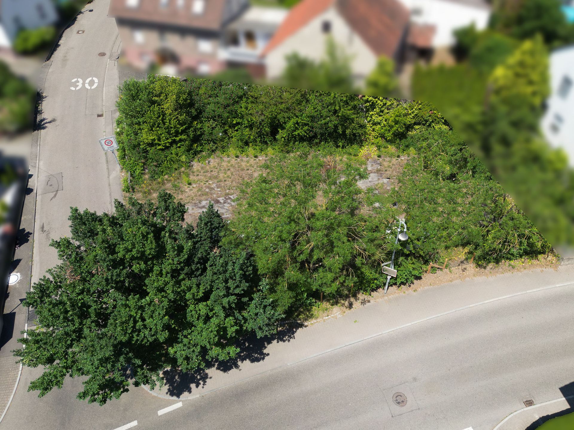 Luftaufnahme eines leeren, überwucherten Grundstücks an einer Straßenecke mit Bäumen.